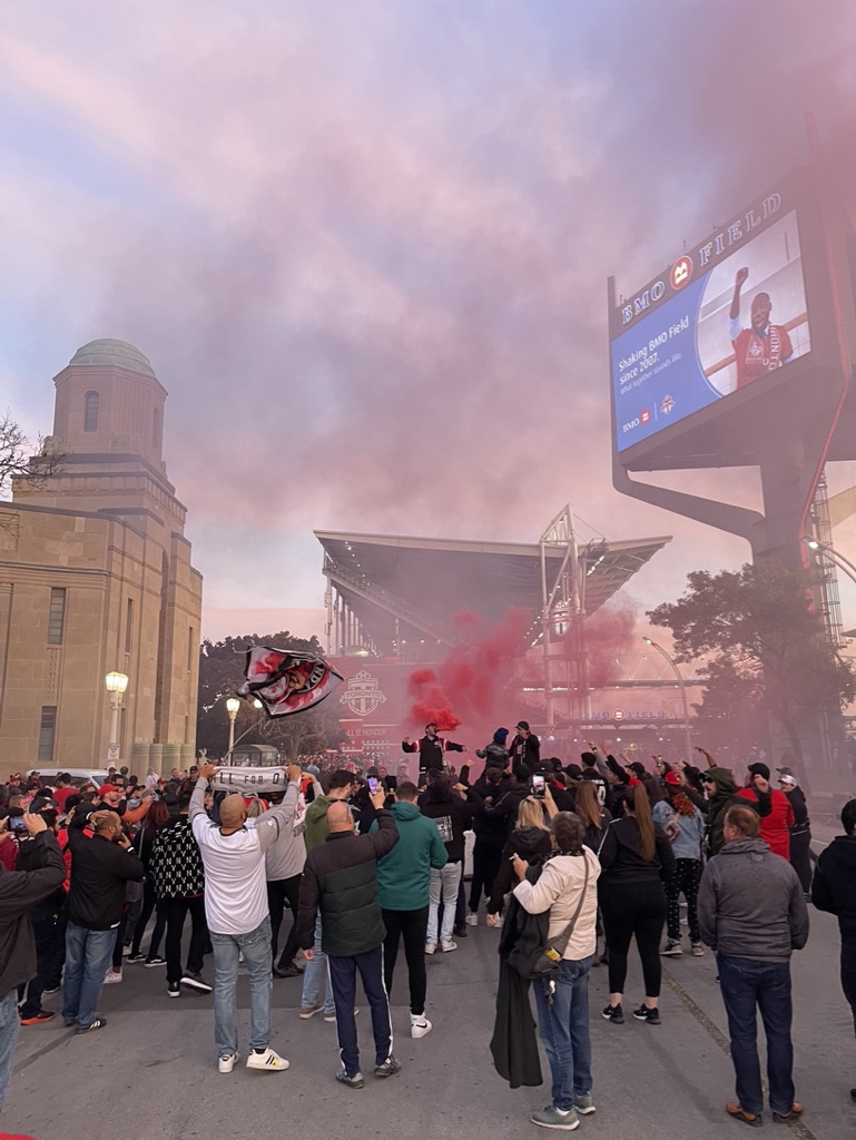 Fans gathered by Toronto BMO field, red smoke in the background in front of screen