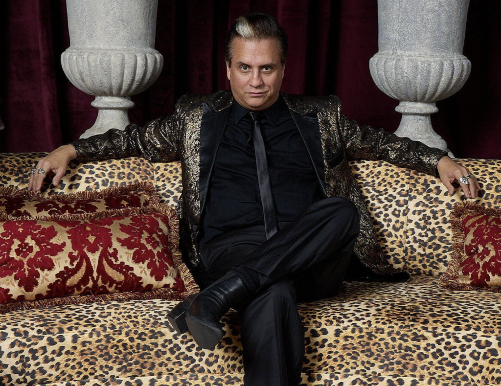 Mysterion the Mindreader Man wearing a suit sitting on a leopard print couch with his arms stretched out.