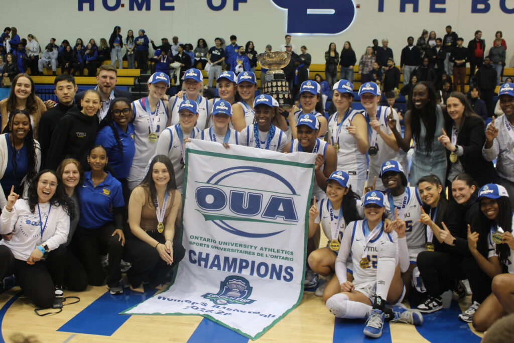 A basketball team poses with a banner and a trophy.