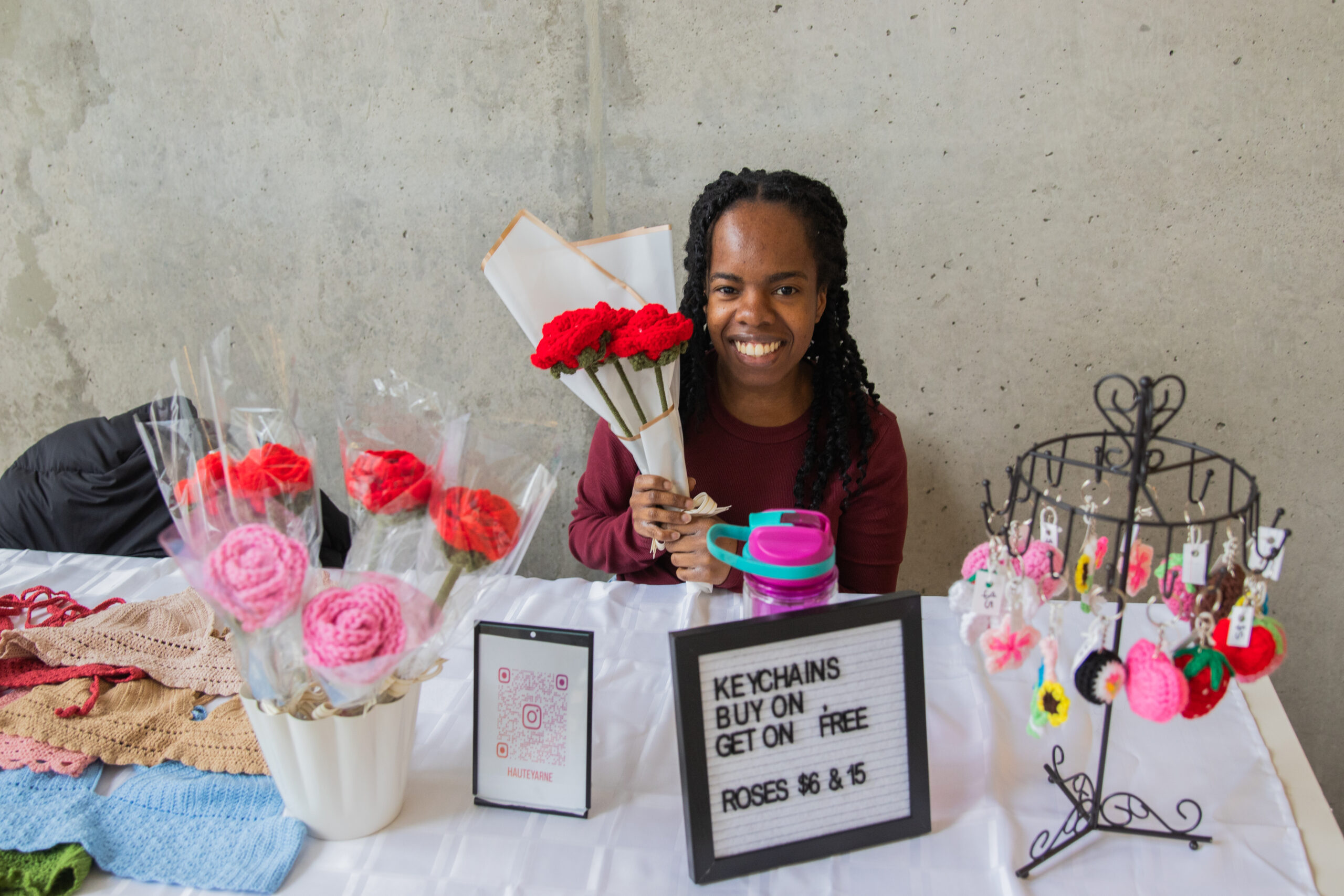 Woman sits in front of white table with crocheted flowers and keychains on it.