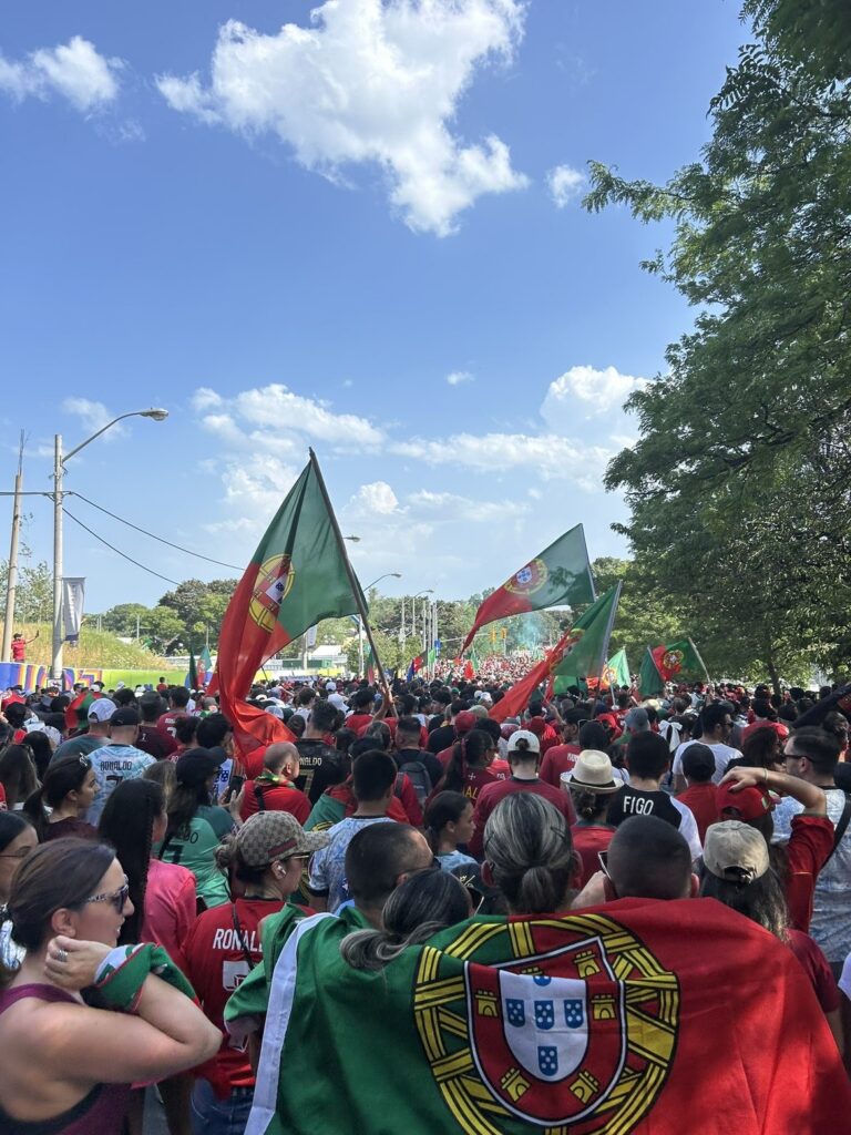large crowd of Portugal soccer fans gathers outdoors, waving Portuguese flags and wearing red and green jerseys under a sunny blue sky.
