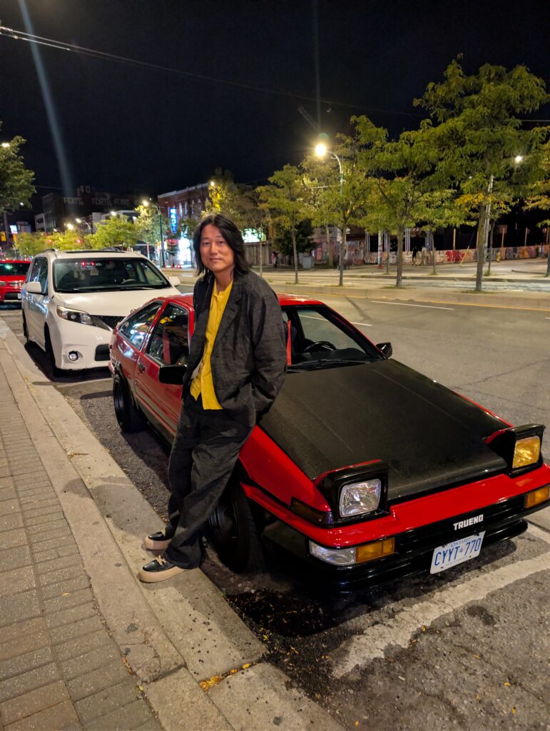 Image of actor Sung Kang leaning against a red car.