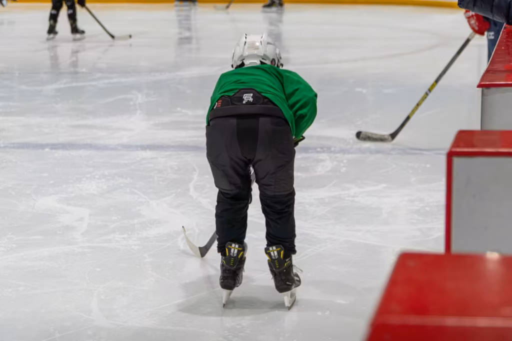 A young hockey player from behind wearing a green jersey and a white helmet, holding a hockey stick while on the ice.