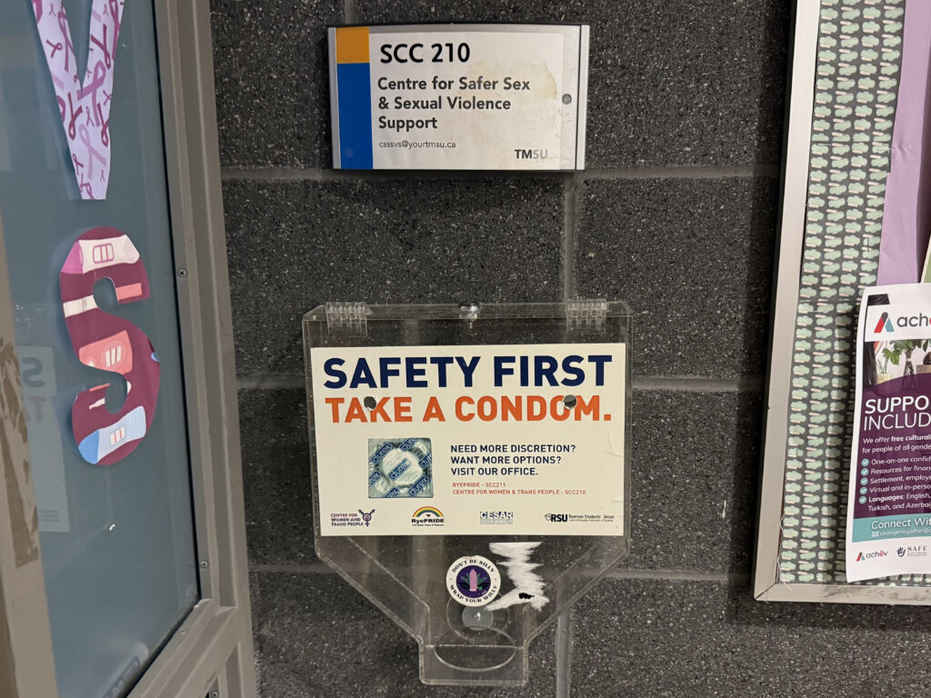 A clear plastic condom dispenser mounted on a dark brick wall at TMU. A sign above identifies the Centre for Safer Sex and Sexual Violence Support, while a sign on the dispenser reads “Safety First, Take a Condom.”
