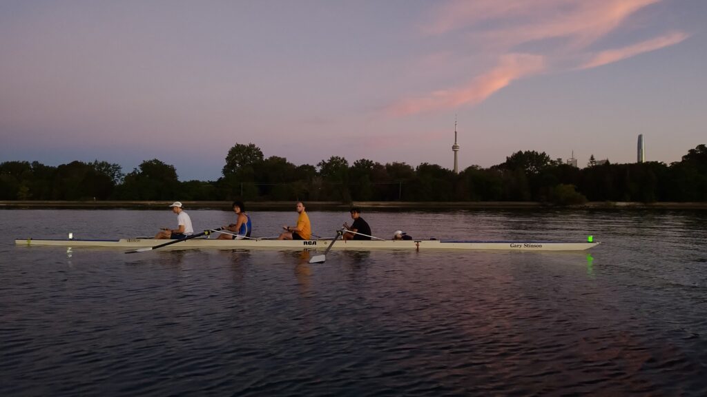 Four rowers sit in a rowing shell on Lake Ontario at sunrise, with Toronto's CN Tower visible in the background