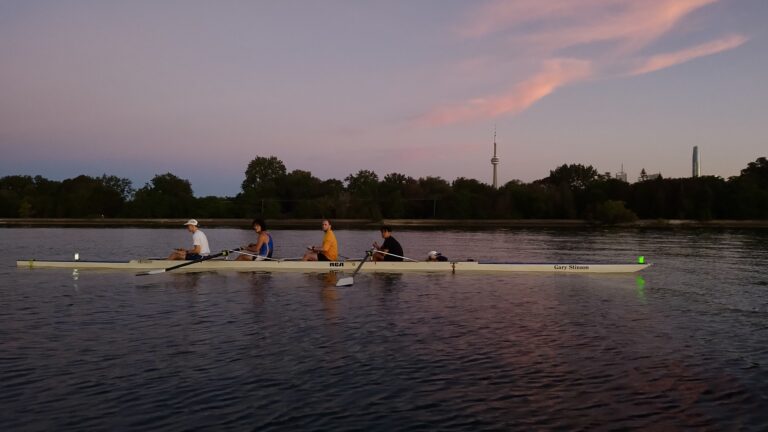 Four rowers sit in a rowing shell on Lake Ontario at sunrise, with Toronto's CN Tower visible in the background