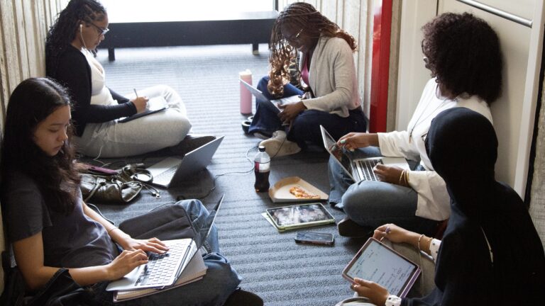 A group of female students doing work on computers, laptops and iPads while sitting in a group.