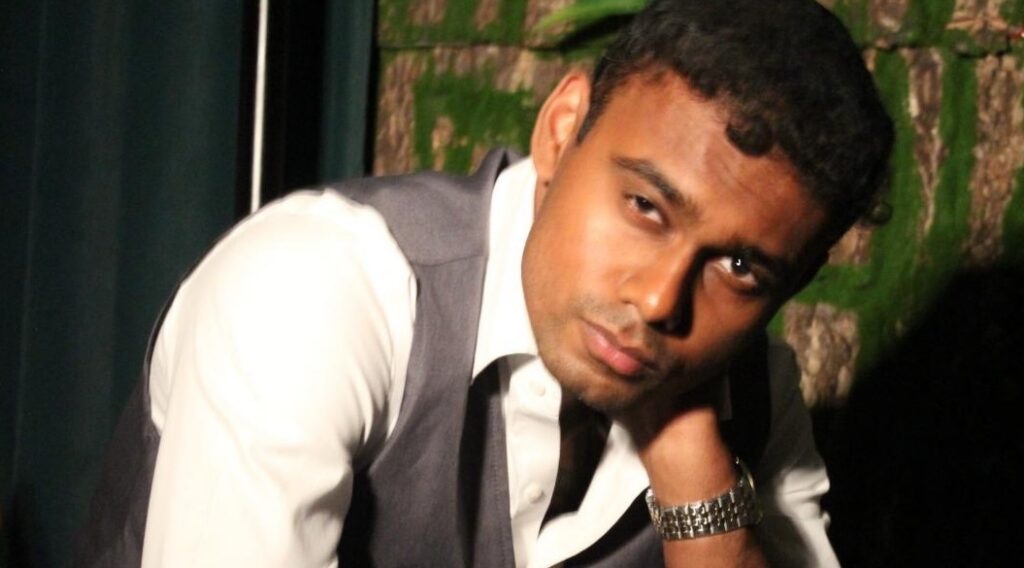 A man in a white shirt and a gray vest is posing with his arm on his chin.