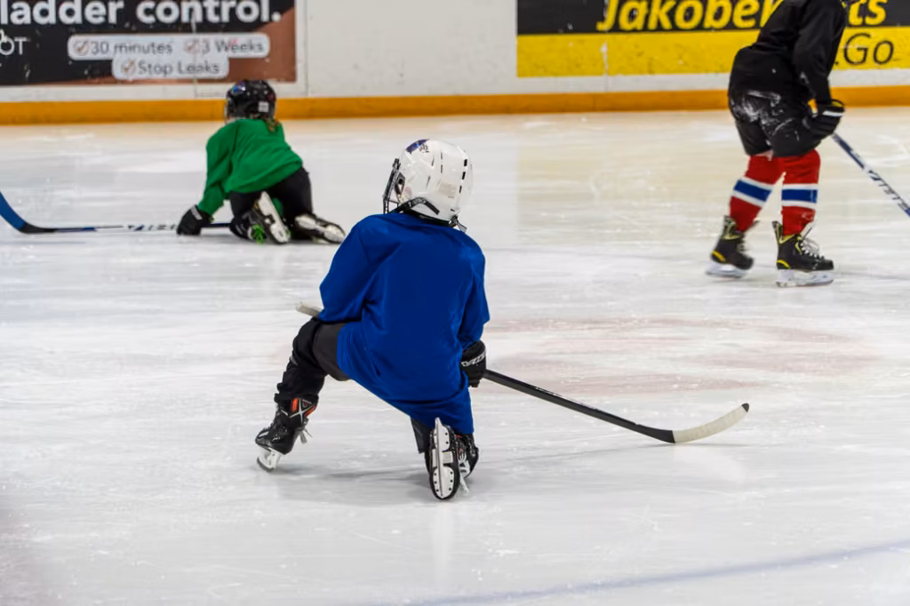 A young hockey player in a blue jersey and white helmet kneels on the ice during practice, holding a hockey stick.
