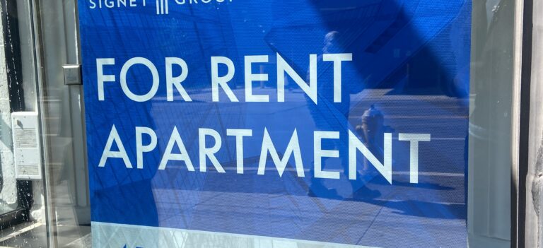 A blue sign in a window reads “FOR RENT APARTMENT” in large white letters