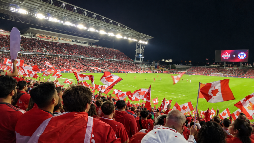 Canadian soccer fans wave Canadian flags and cheer from the stands during a match at a packed stadium at night.