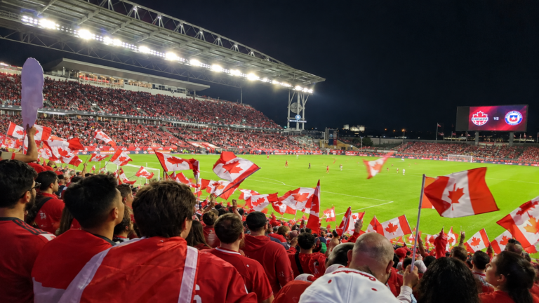 Canadian soccer fans wave Canadian flags and cheer from the stands during a match at a packed stadium at night.