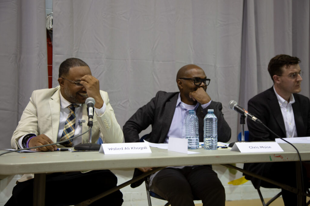 Three men sitting at tables on chairs, two are laughing and smiling.