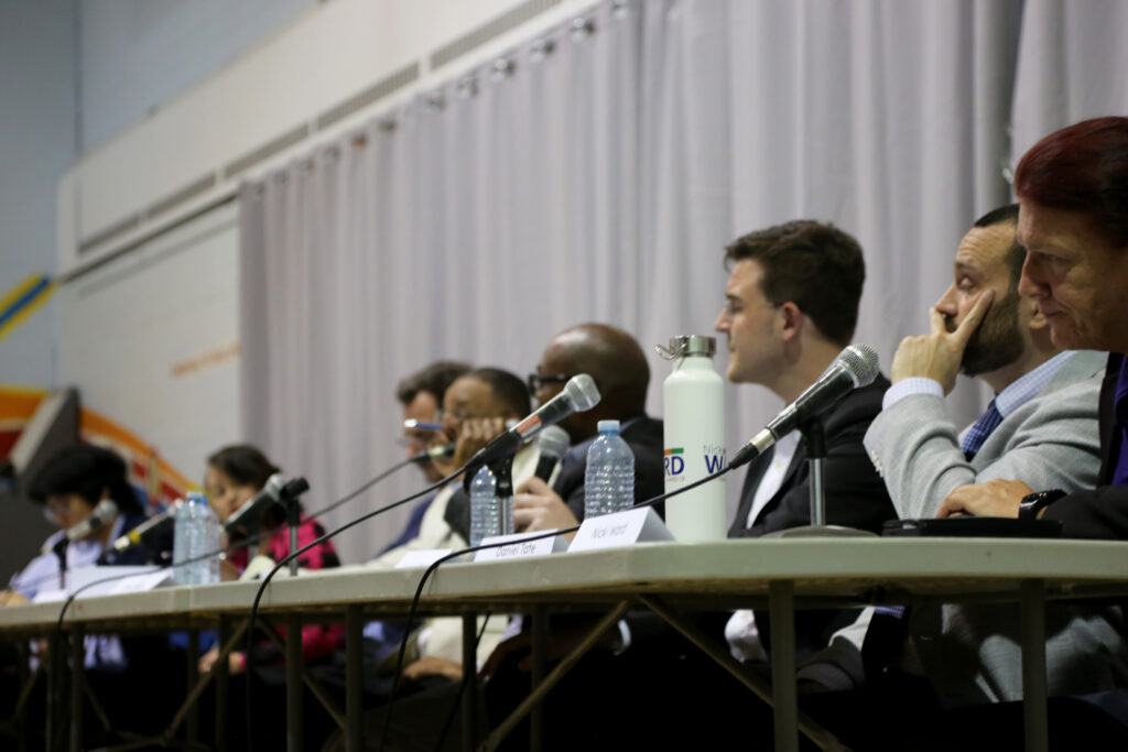 People sit at tables on chairs in a line with mics.