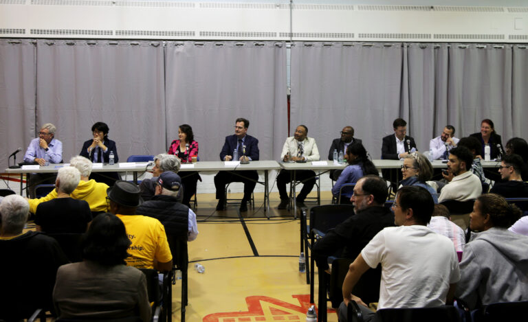 Nine people sit at tables on chairs during a municipal election debate as audience members watch from rows of chairs in front of them.