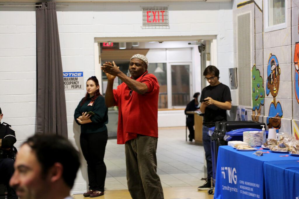 A man wearing a red t-shirt and brown pants and a head covering standing up and clapping.