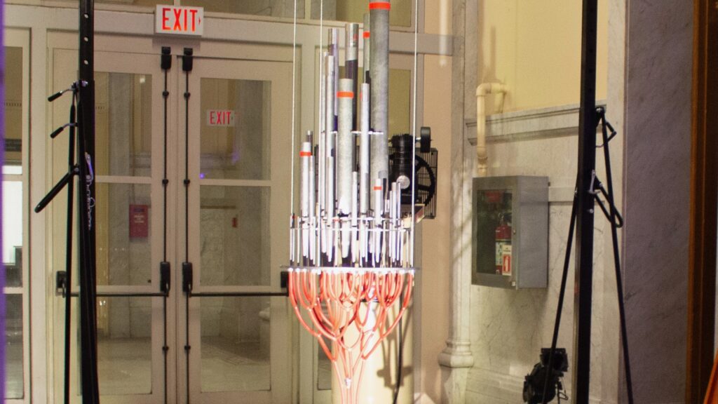 A bunch of church organs being held up by wires glowing from lighting in front of an exit signed door.