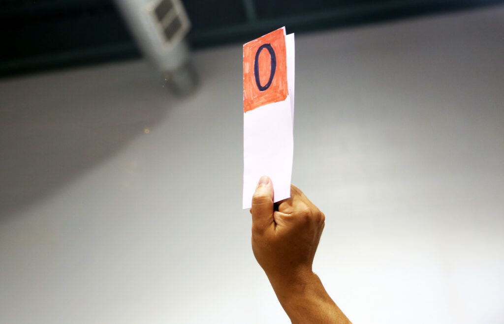 ALT TEXT: A hand holding up a paper with a red square with the number zero.