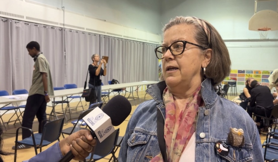 A woman with short grey hair wearing a headband, a blue jean jacket and a pink scarf looking to the left, talking into a microphone.