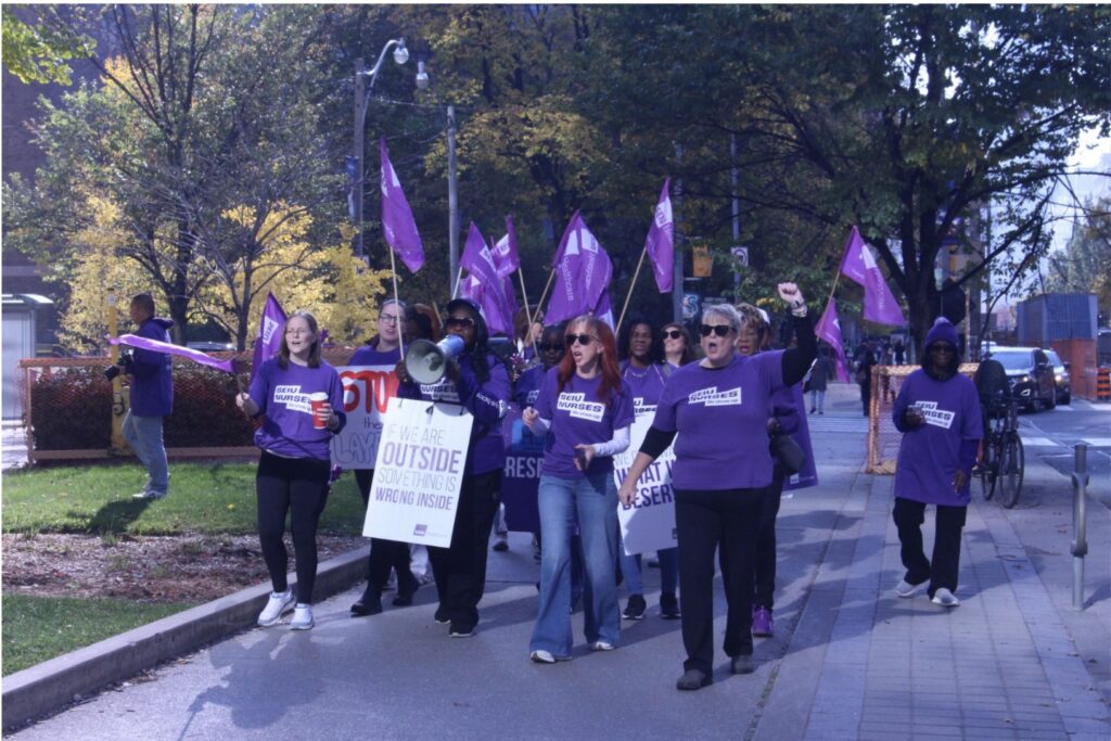 A group of people wearing matching purple shirts that say “SEIU Nurses: we show up” flags and signs on a sidewalk.