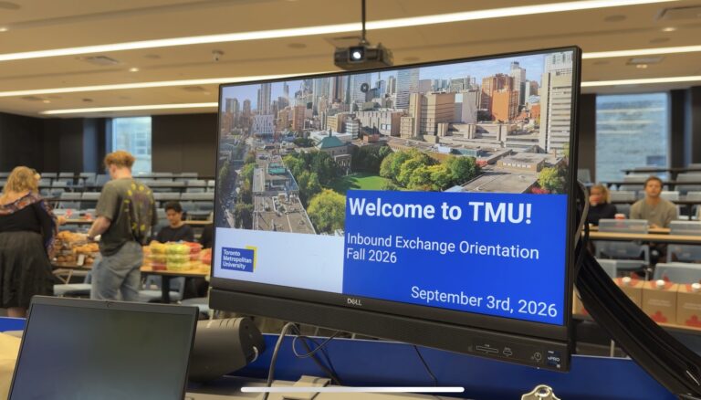 A monitor screen displays 'Welcome to TMU! Inbound Exchange Orientation Fall 2026' with two students in the background.