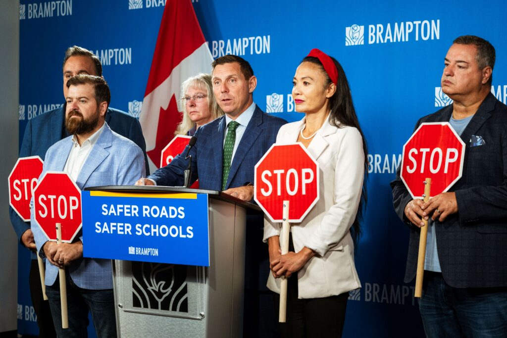 People hold red stop signs beside a speaker at a podium marked “Safer Roads Safer Schools,” against a blue Brampton backdrop.