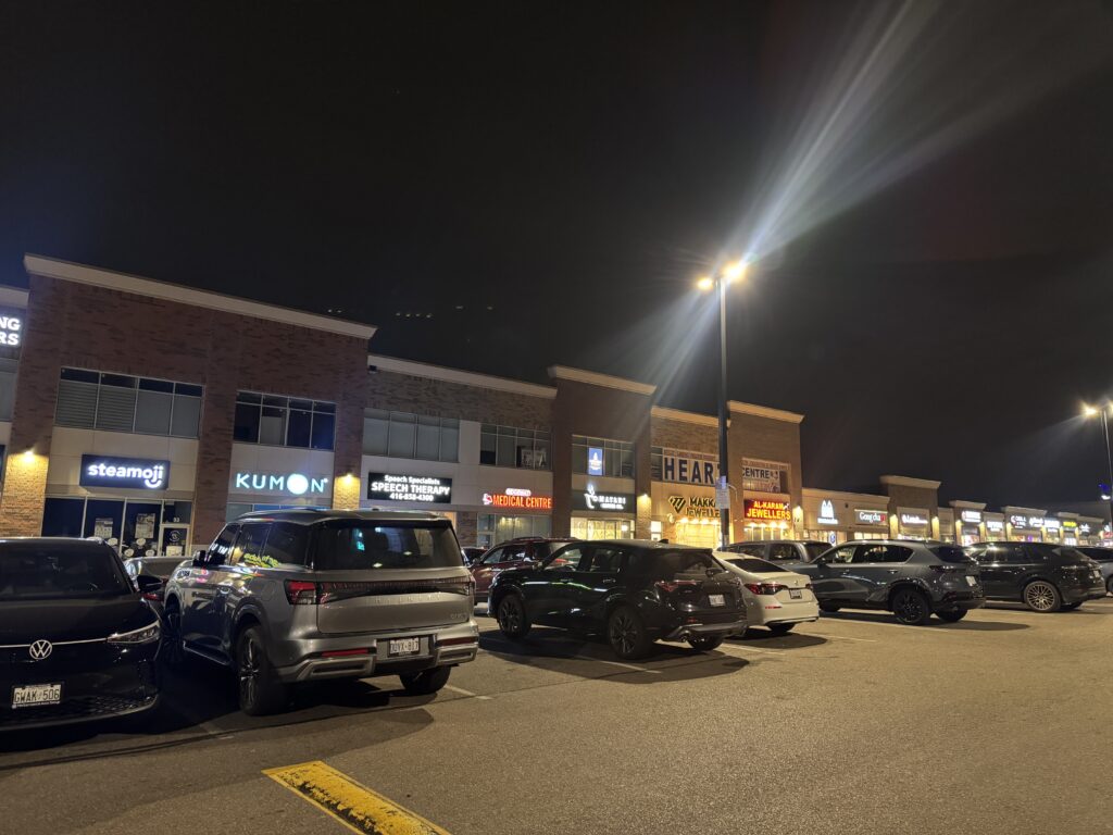 Night time at Ridgeway Plaza, with different storefronts in the background and cars parked in front of them