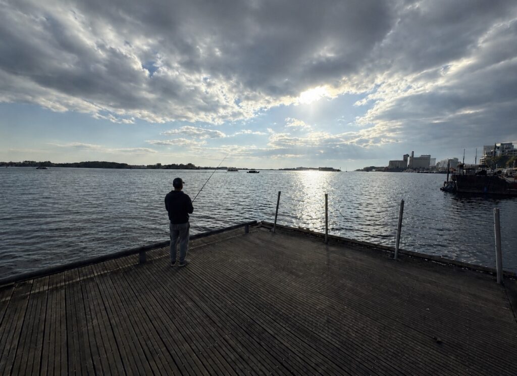 A lone angler stands on a wooden pier near Toronto’s Harbourfront Centre, casting a fishing rod into Lake Ontario beneath a cloudy sky with sunlight reflecting off the water.