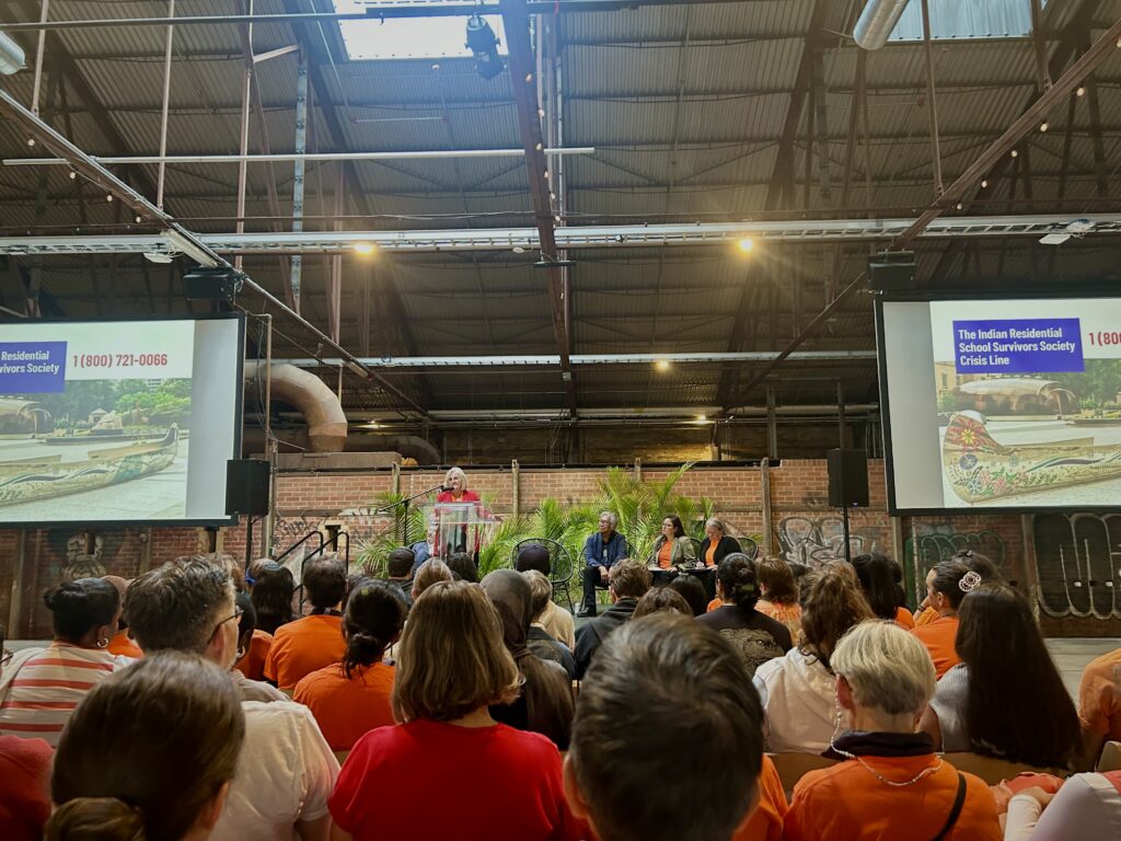 Large room with an audience wearing orange shirts and a stage with speakers presenting.